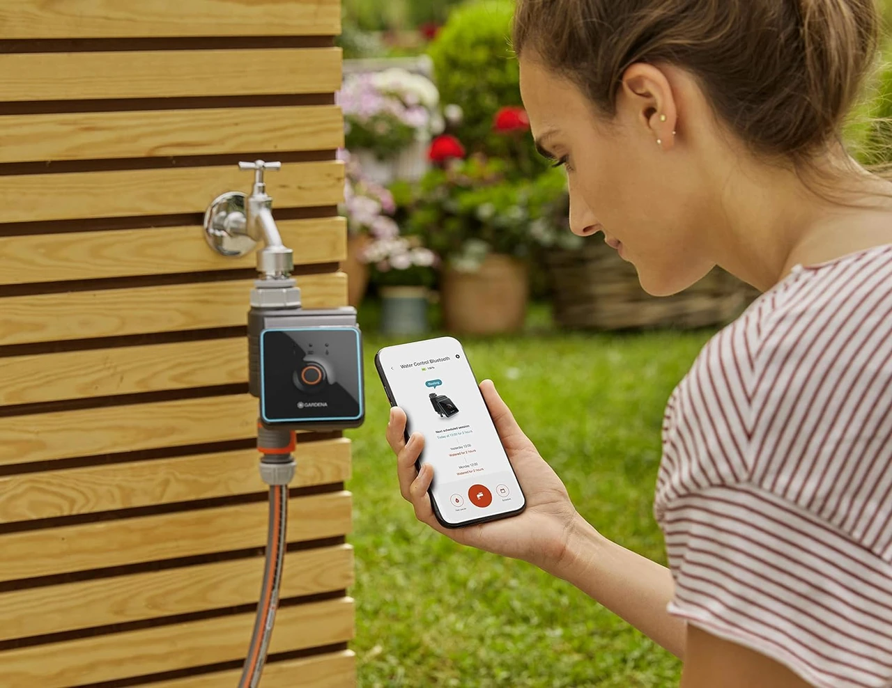 Eine Frau bedient ein automatisches Bewässerungssystem im Garten mit einer Smartphone-App. Der Garten im Hintergrund zeigt gesunde Pflanzen.