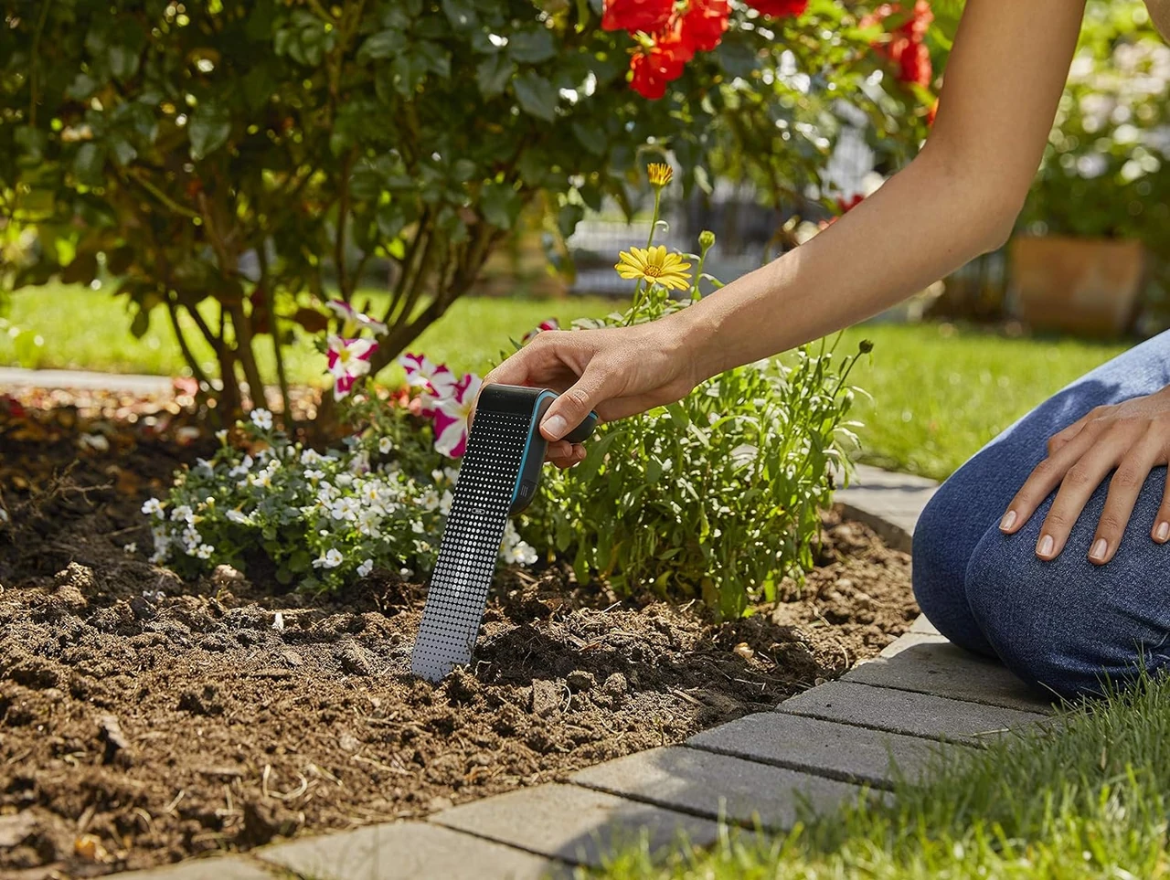 Eine Person platziert einen Gardena Feuchtigkeitssensor in den Boden eines Gartens, um die Bodenfeuchtigkeit zu messen und die automatische Bewässerung zu optimieren.