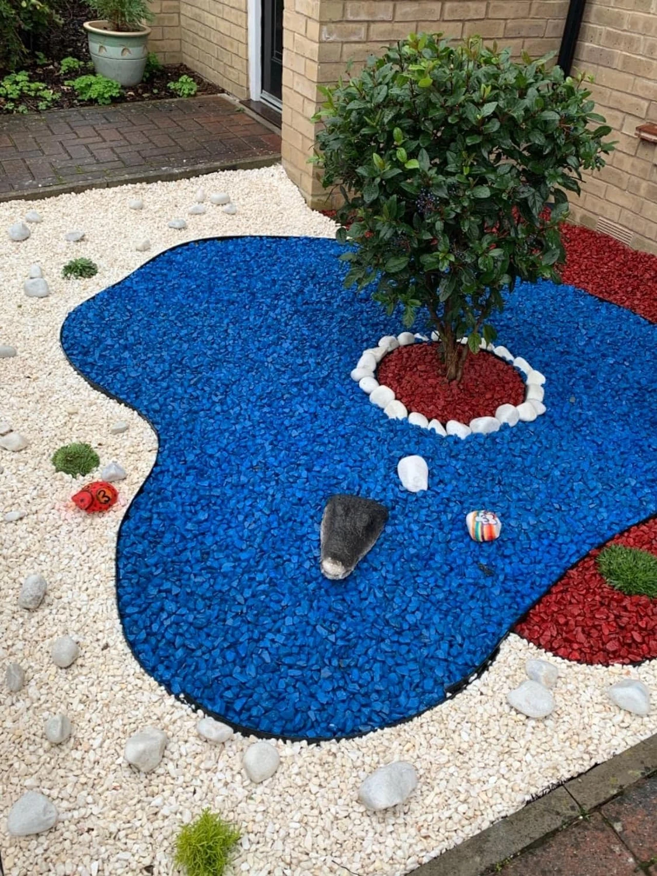 Ein farbenfrohes Kiesbeet mit blauem, rotem und weißem Gartenkies, das um einen kleinen Baum herum angelegt ist. Perfekt für die Gestaltung eines Steinbeets im Garten.